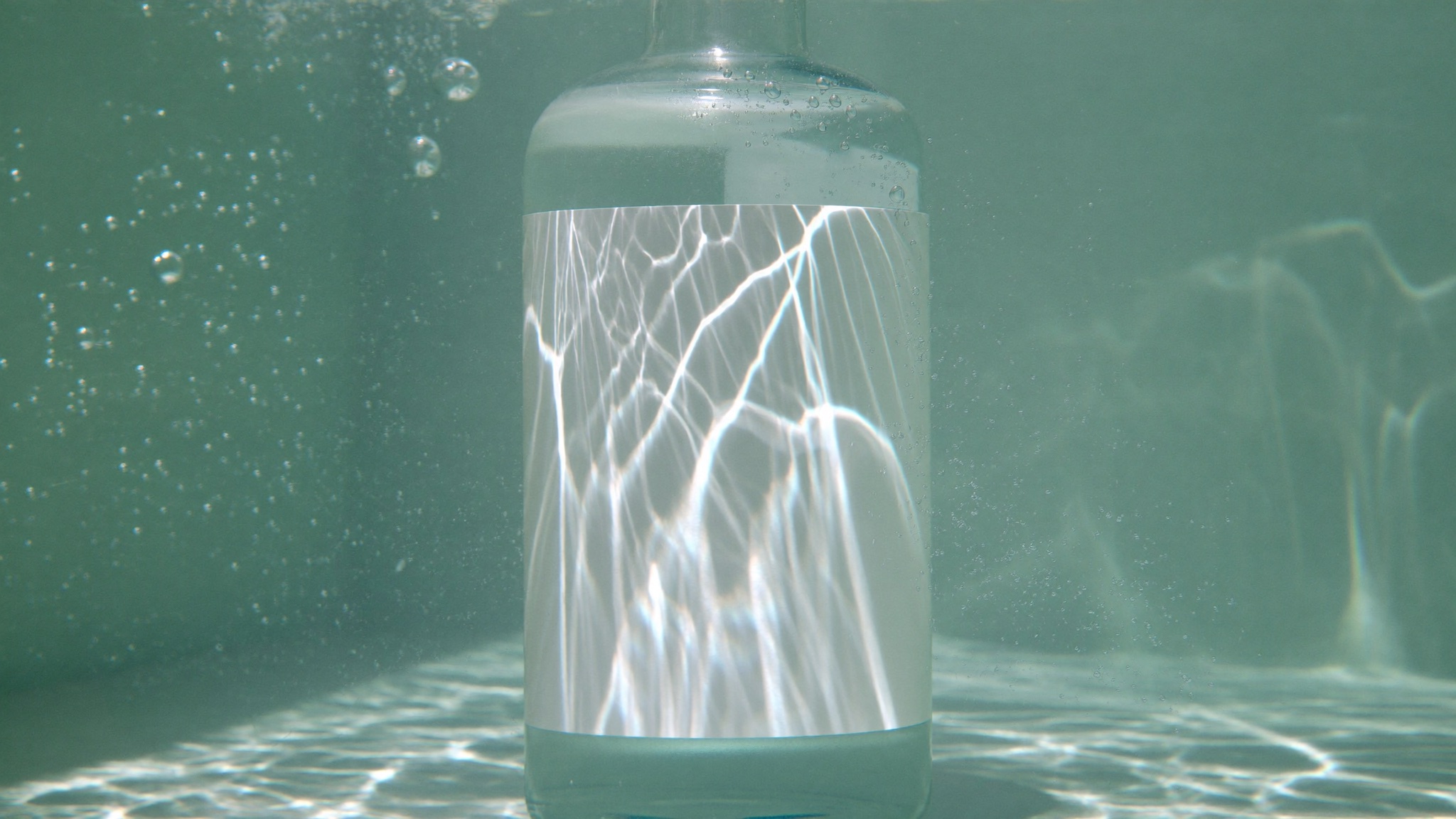 A frosted glass bottle suspended underwater, white caustic light raking across its blank label