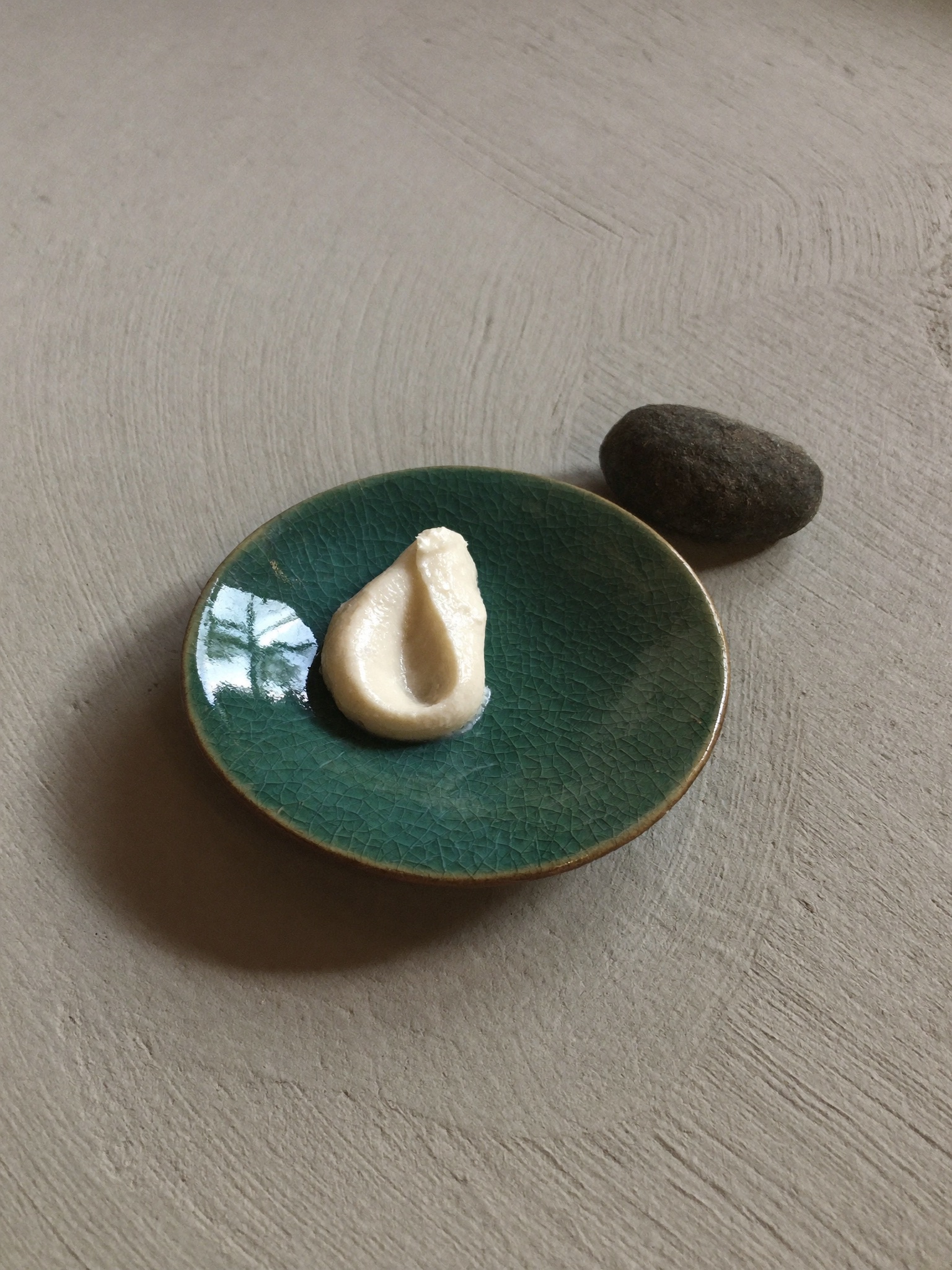 A celadon dish with crackle glaze holding a swirl of white cream, beside a river stone
